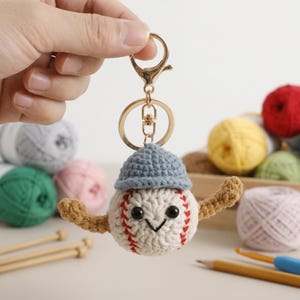 May include: A handmade crochet baseball keychain with a smiling face, red stitching, and a blue cap. The keychain has a gold-colored clasp and ring. The baseball is holding a brown crochet bat. The background includes yarn balls and knitting needles.