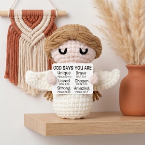 May include: A crocheted angel doll with a light brown halo and hair, holding a sign that reads "GOD SAYS YOU ARE Unique Brave Loved Chosen Strong Amazing" with Bible verses. The doll is on a wooden shelf.