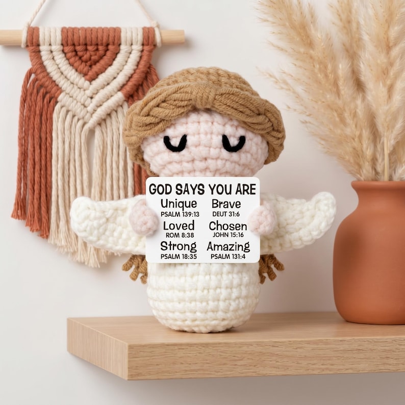 May include: A crocheted angel doll with a light brown halo and hair, holding a sign that reads "GOD SAYS YOU ARE Unique Brave Loved Chosen Strong Amazing" with Bible verses. The doll is on a wooden shelf.