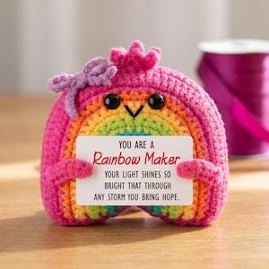 May include: A crocheted rainbow-colored plush toy with a pink top, a purple bow, and black bead eyes. It holds a white rectangular card that reads: YOU ARE A Rainbow Maker YOUR LIGHT SHINES SO BRIGHT THAT THROUGH ANY STORM YOU BRING HOPE.