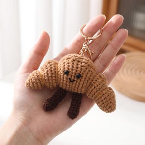 May include: A light brown crocheted croissant keyring featuring two small black plastic eyes and a stitched black smile. The croissant has two dark brown dangling legs and is attached to a gold-toned metal split ring and lobster clasp.