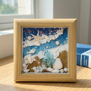 Könnte beinhalten: Ein gerahmtes Kunstwerk mit Meeresmotiv, das einen Seestern, Muscheln, Perlen und eine Miniaturflasche zeigt. Das Kunstwerk verwendet Sand, blauen Glitzer und andere Materialien. Der Rahmen ist aus hellem Holz gefertigt.