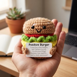 May include: A crocheted burger with a smiling face, red tomato, green lettuce, and yellow cheese. The burger holds a sign that reads "Positive Burger" with an inspirational message. A fun, handmade item.