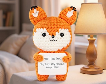 Puede incluir: Un peluche de zorro tejido a ganchillo en color naranja y blanco con ojos y orejas negros, que sostiene un cartel rectangular blanco que dice: Positive fox. Stay foxy, stay fabulous. You got this! El zorro tiene una cabeza redondeada.