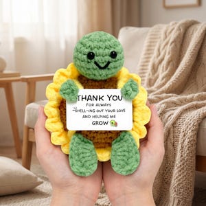 Puede incluir: Una tortuga de ganchillo hecha a mano con un cuerpo de girasol. La tortuga es verde con una cara sonriente y sostiene un letrero que dice "THANK YOU FOR ALWAYS SHELL-ING OUT YOUR LOVE AND HELPING ME GROW."