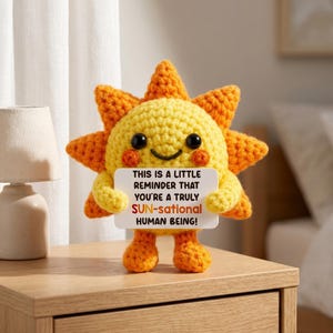 Może przedstawiać: Szydełkowa figurka w kształcie słońca z uśmiechniętą twarzą, trzymająca znak z napisem "THIS IS A LITTLE REMINDER THAT YOU'RE A TRULY SUN-sational HUMAN BEING!". Słońce jest żółte i pomarańczowe.