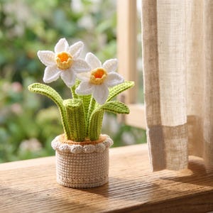 Könnte beinhalten: Eine handgefertigte Häkel-Topfpflanze mit zwei weißen Narzissenblüten mit leuchtend gelben Zentren. Die Blumen haben grüne Stängel und Blätter und stehen in einem strukturierten, cremefarbenen, zylindrischen Häkeltopf mit einem dekorativen Rand.