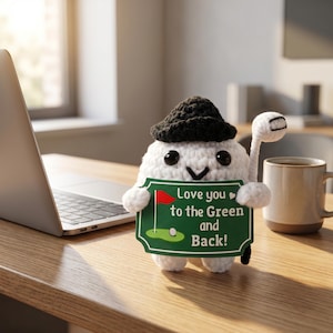May include: A white crocheted golf ball character wearing a black hat, holding a golf club and a green sign that reads "Love you to the Green and Back!" The sign features a golf course illustration.