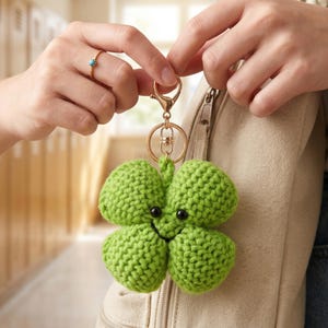 May include: A green crocheted four-leaf clover keychain with a gold-colored clasp and ring. The clover has a smiling face with black eyes. The keychain is held by a person.