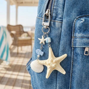 May include: A decorative keychain with a beach theme, featuring a large off-white starfish, a white seashell, and a small blue fish charm. The keychain is attached to a blue denim backpack with a silver-colored clasp.