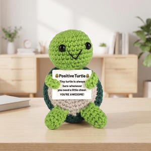 May include: A crocheted green turtle figurine holding a sign that reads "Positive Turtle." The sign continues with the text "Tiny turtle is always here whenever you need a little cheer. YOU'RE AWESOME!" The turtle has black eyes and a smiling face.
