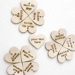 May include: Wooden heart-shaped ornaments with engraved messages. Some are arranged in four-leaf clover shapes, with phrases like "Mom My Star" and "Happy Birthday." One heart reads "your text here."