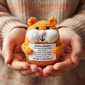 May include: A small, round, orange and white crocheted hamster toy with black eyes and a pink nose. It holds a white rectangular card with the text: Positive Hamster, Life's a maze, but you're doing wheel-y great! Keep nibbling at your dreams.