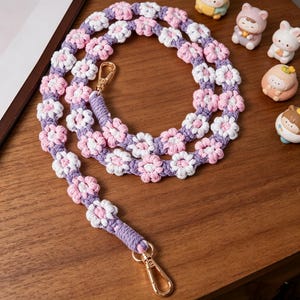 May include: A handmade macrame strap featuring a floral design in white, pink, and purple. The strap has gold-colored metal clasps on each end. The strap is lying on a wooden surface.