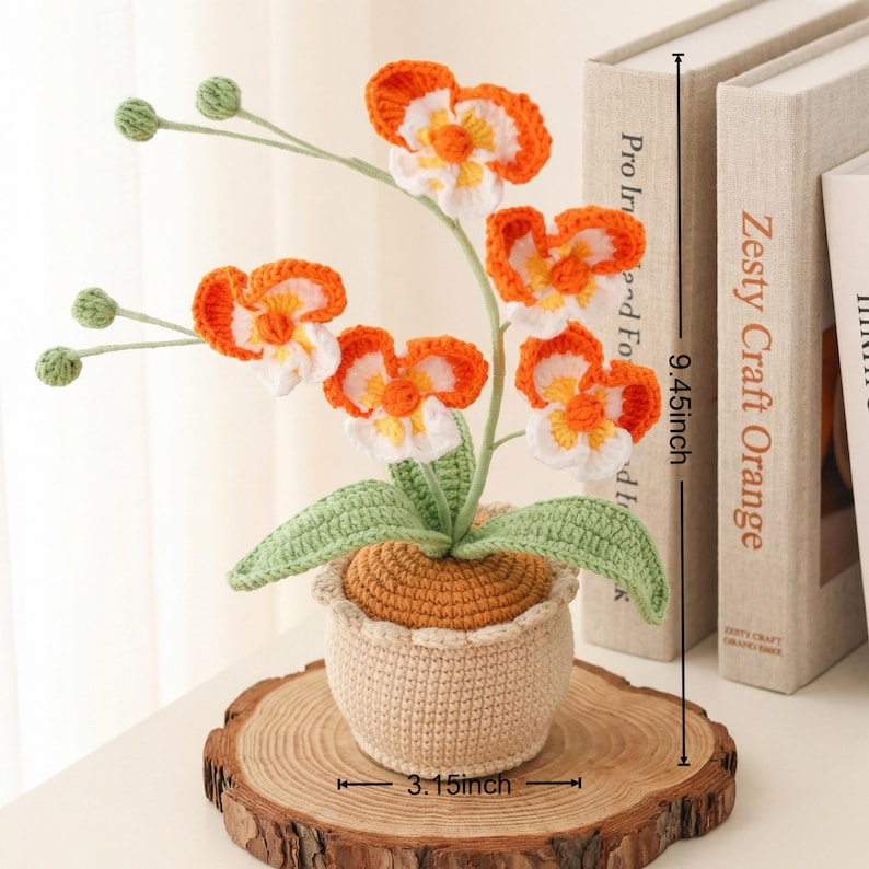 May include: A crocheted orchid plant with orange and white flowers, green leaves, and a beige pot. The plant is 9.45 inches tall and 3.15 inches wide, sitting on a wooden slice.