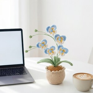 May include: A crocheted orchid plant in a beige pot. The flowers are blue, white, and yellow, with green stems and leaves. The pot has a scalloped edge. A laptop and a cup of coffee are in the background.