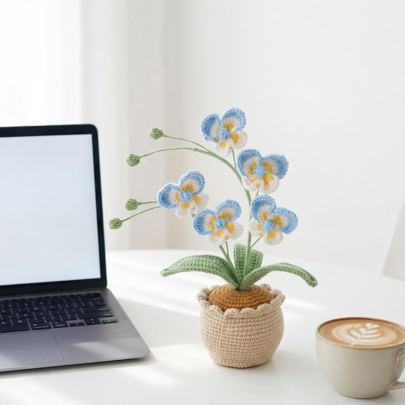 May include: A crocheted orchid plant in a beige pot. The flowers are blue, white, and yellow, with green stems and leaves. The pot has a scalloped edge. A laptop and a cup of coffee are in the background.
