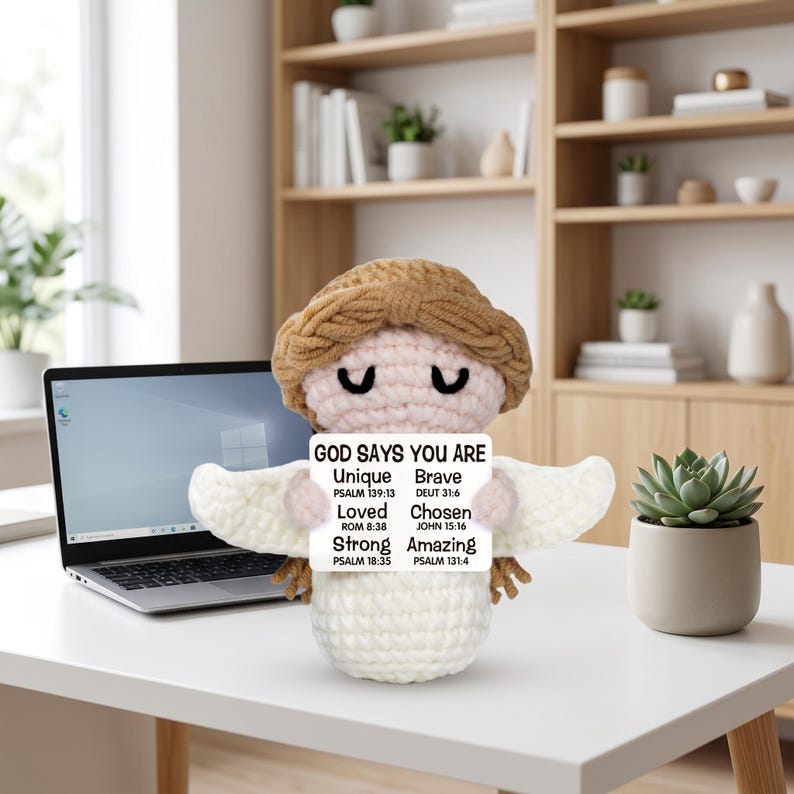 May include: A handmade crochet angel doll with a light brown halo and hair, holding a sign that reads "GOD SAYS YOU ARE Unique Brave Loved Chosen Strong Amazing". The angel has white wings and is sitting on a white desk.