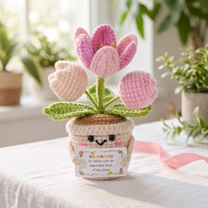 Könnte beinhalten: Eine gehäkelte Topfpflanze mit drei rosa und hellrosa Tulpenblüten, grünen Stielen und Blättern. Der beige Topf hat ein lächelndes Gesicht mit schwarzen Augen und rosa Wangen und hält eine weiße Karte mit der Aufschrift, THANK YOU for being such an important part of my story.