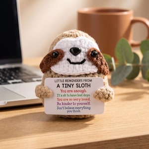 May include: A crocheted tan and white sloth figurine holding a white rectangular card that reads: LITTLE REMINDERS FROM A TINY SLOTH. You are enough. It's ok to have bad days. You are so very loved. Be kinder to yourself. Don't believe everything you think.