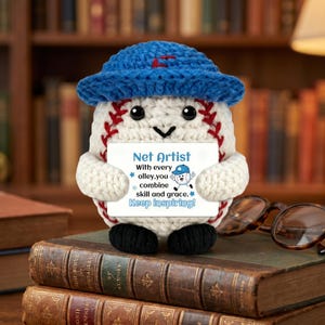 May include: A crocheted baseball character wearing a blue hat, holding a sign that reads "Net Artist." The baseball has red stitching and black eyes. The sign has text that says "With every olley, you combine skill and grace. Keep inspiring!"
