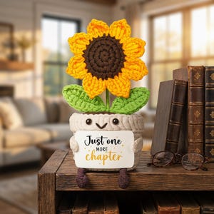 Pode incluir: Um girassol de crochê em um vaso com um rosto sorridente, segurando uma placa que diz "Just one more chapter". O girassol tem pétalas amarelas e um centro marrom. O vaso é bege e as folhas são verdes. O item está em uma prateleira de madeira com livros.