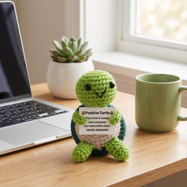 May include: A handmade green and blue crocheted turtle figurine holding a sign that reads "Positive Turtle". The sign says "Tiny turtle is always here whenever you need a little cheer. YOU'RE AWESOME!" A green mug and a laptop are in the background.