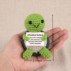 May include: A crocheted green turtle with a smiling face and black eyes. The turtle holds a sign that reads "Positive Turtle" and a message of encouragement. The turtle measures 3.93 inches tall and 2.75 inches wide.