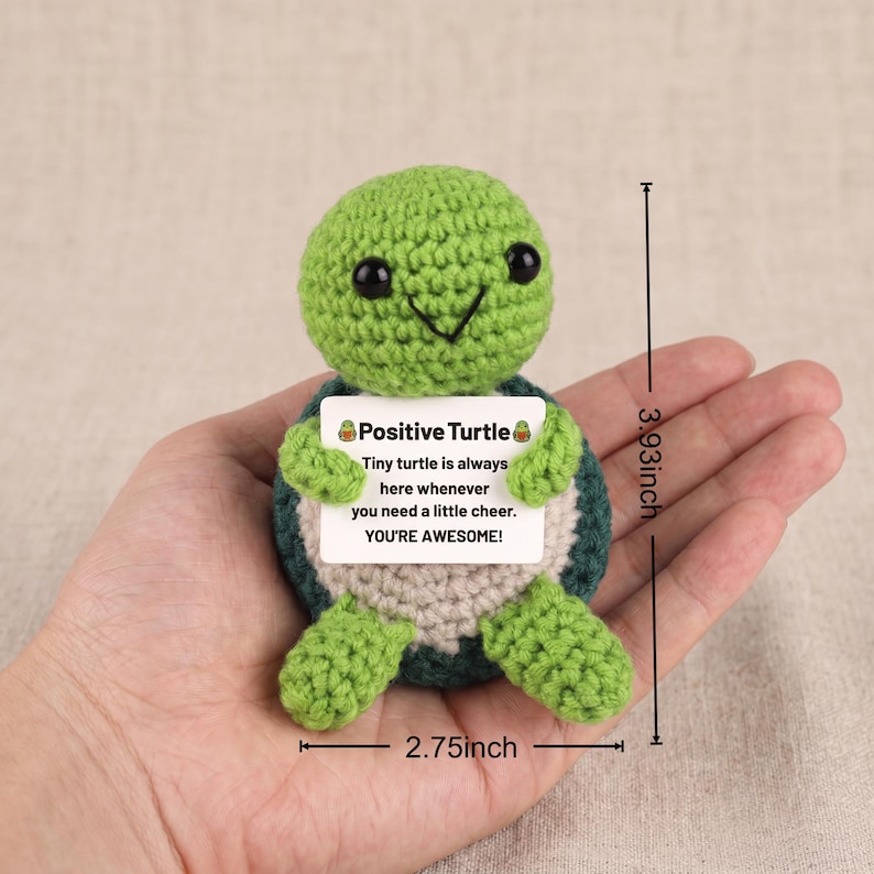 May include: A crocheted green turtle with a smiling face and black eyes. The turtle holds a sign that reads "Positive Turtle" and a message of encouragement. The turtle measures 3.93 inches tall and 2.75 inches wide.