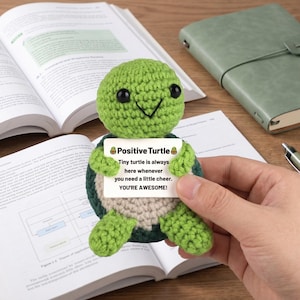 May include: A crocheted green turtle holding a sign that reads "Positive Turtle." The sign says "Tiny turtle is always here whenever you need a little cheer. YOU'RE AWESOME!" The turtle is on a wooden surface with books and a notebook.