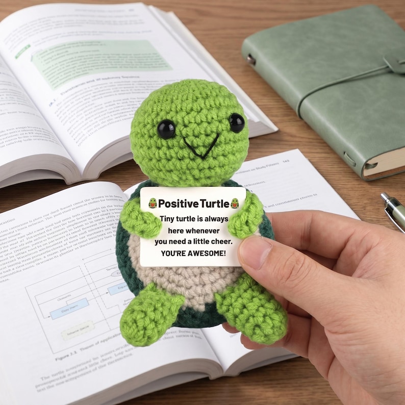 May include: A crocheted green turtle holding a sign that reads "Positive Turtle." The sign says "Tiny turtle is always here whenever you need a little cheer. YOU'RE AWESOME!" The turtle is on a wooden surface with books and a notebook.