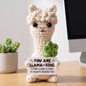 May include: A cream-coloured crocheted llama figurine featuring a textured wool-like surface, black plastic eyes, pink cheeks, and a green crocheted four-leaf clover. It holds a white sign that reads: YOU ARE LLAMA-ZING A TINY LLAMA IS HERE TO ALWAYS REMIND YOU.