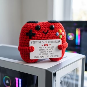 May include: A red crocheted plush toy shaped like a game controller with black button eyes and a smiling mouth. It holds a white card that reads: POSITIVE GAME CONTROLLER. I MAY BE A TINY GAME CONTROLLER BUT I BELIEVE IN YOU. GO DO YOUR THING! YOU'RE NEXT LEVEL AWESOME!