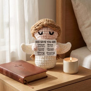 May include: A crocheted angel doll with a sign that reads "GOD SAYS YOU ARE Unique Brave Loved Chosen Strong Amazing". A brown leather-bound Holy Bible and a lit candle sit on a wooden nightstand.