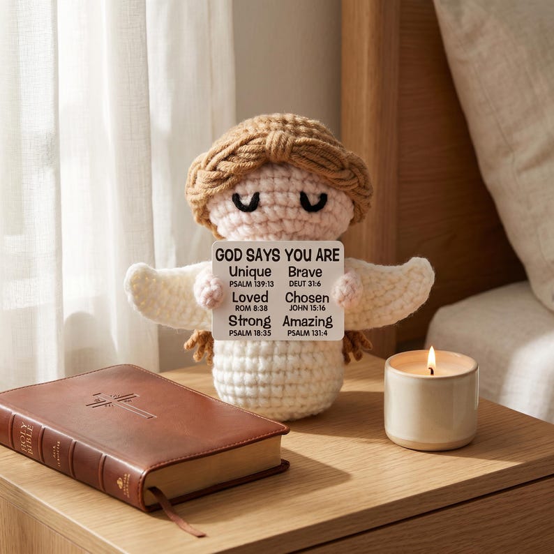 May include: A crocheted angel doll with a sign that reads "GOD SAYS YOU ARE Unique Brave Loved Chosen Strong Amazing". A brown leather-bound Holy Bible and a lit candle sit on a wooden nightstand.
