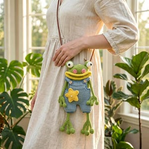 May include: A crochet frog-shaped crossbody bag featuring a green frog with large white eyes, pink cheeks, and a wide yellow mouth. The frog wears light blue crochet overalls with two small brown buttons and a yellow star applique on the front.