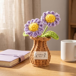 May include: A crocheted tan vase containing two purple flowers with bright yellow centers and green leaves. The vase features a white fabric label on the front with the text HandMade with love and decorative branch illustrations on each side.