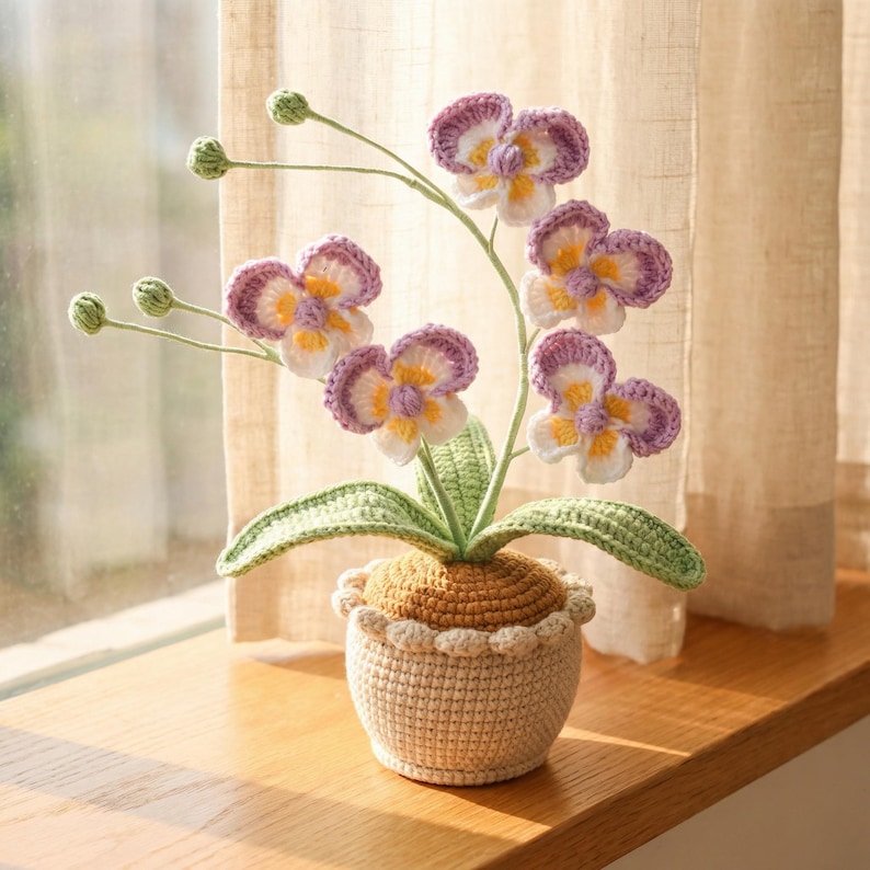May include: A crocheted orchid plant in a beige pot. The orchid features purple, white, and yellow flowers, green leaves, and stems. The pot has a scalloped edge and sits on a wooden surface. The background includes a sheer curtain.