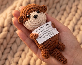 May include: A small, brown and cream-colored crocheted otter toy holding a white rectangular card. The card features black text that reads: Positive Otter, I may be a tiny otter, but I believe in you. Keep your head up & float on. YOU'RE OTTERLY AMAZING!
