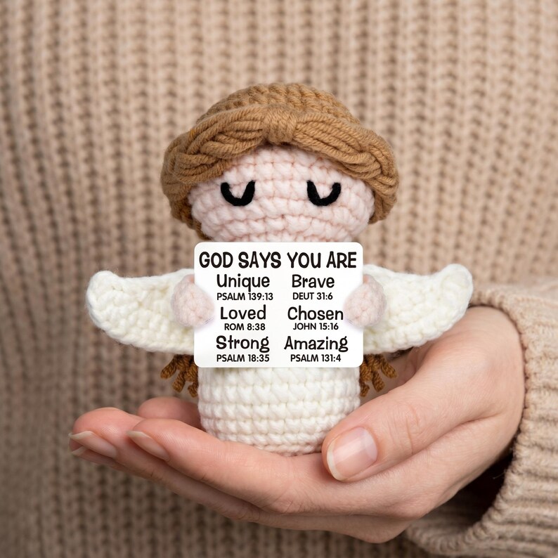 May include: A crocheted angel doll with a brown halo and hair, white wings, and a white body. The doll holds a sign that reads "GOD SAYS YOU ARE Unique, Brave, Loved, Chosen, Strong, Amazing" with Bible verses.