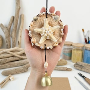 Puede incluir: Un carillón hecho a mano con una rodaja de madera adornada con conchas marinas, perlas y una estrella de mar. Una campana dorada cuelga debajo. El carillón está suspendido por un cordón negro, creando un tema costero o náutico.