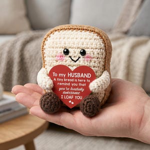 May include: A small, crocheted plush toy shaped like a slice of bread with a tan crust and cream-colored center. It features black eyes, pink cheeks, and a smile. It holds a red heart-shaped card with the text: To my HUSBAND A tiny bread is here to remind you that you're toastally awesome I LOAF YOU.