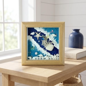 May include: A framed beach scene with a light wood frame. The artwork features a blue and white ocean design with seashells, pearls, and a starfish. The frame is square and sits on a wooden table.