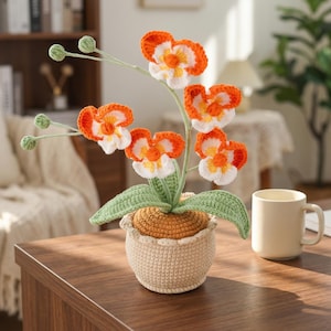 Op de afbeelding: Een handgemaakte gehaakte orchideeplant in een beige pot. De bloemen zijn oranje en wit met gele centra. Groene bladeren en stelen ondersteunen de bloemen. De pot staat op een bruine houten tafel naast een beige mok.