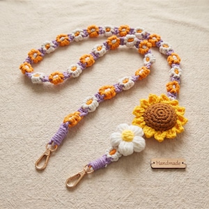 May include: A crochet lanyard featuring a repeating pattern of small orange and white flowers on a purple cord. The end includes a large yellow sunflower, a white daisy, and two gold-tone metal lobster clasps. A small wooden tag reads Handmade.