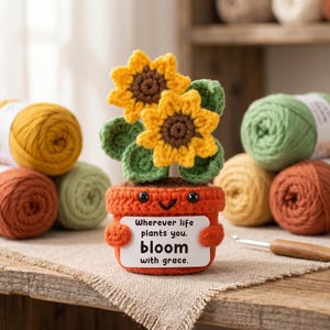 May include: A crocheted orange flower pot with a smiling face, holding a sign that reads "Wherever life plants you, bloom with grace." The pot contains two yellow sunflowers with brown centers and green leaves. Yarn balls in various colors surround the pot.