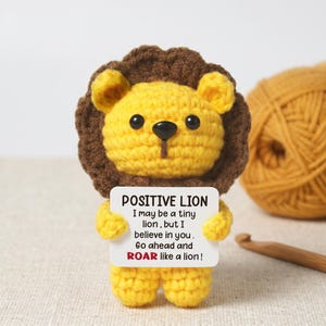 Puede incluir: Un león de ganchillo hecho a mano, amarillo con una melena marrón, sosteniendo un letrero con la inscripción "POSITIVE LION" y un mensaje motivador. Una madeja de lana y un ganchillo son visibles en el fondo.