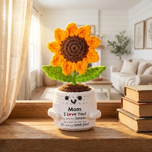 May include: A handcrafted crochet sunflower in a white pot with a smiling face and the text "Mom I Love You! You are my Sunshine. No matter how old I get, I'll always need you!" The sunflower has yellow petals, a brown center, and green leaves.