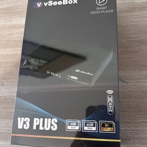 Op de afbeelding: Een zwarte V3 PLUS smart media player box met het vSeeBox logo. De doos heeft de tekst "V3 PLUS", "4GB RAM", "64GB ROM" en "8K ULTRA HD". Een afstandsbedieningspictogram is ook zichtbaar.
