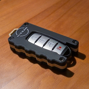 May include: A black and gray Nissan car key fob with silver buttons. The key fob has a ribbed design and the Nissan logo is visible. The key fob is lying on a wooden surface.